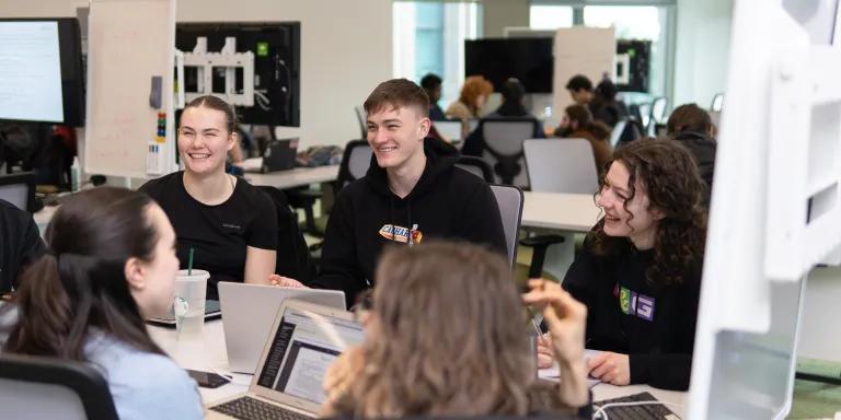 A group of students gather around a table working on their laptops.