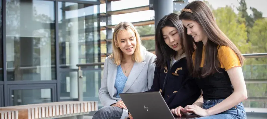Three students sit around a laptop, working together and smiling.